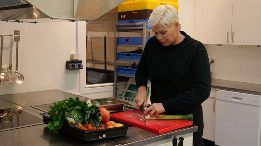 Der ses en kvindelig værkmester. Hun står i et køkken og er i gang med at skære en porre ud på et spækbræt. På køkkenbordet foran værkmesteren står en bakke med grøntsager. 